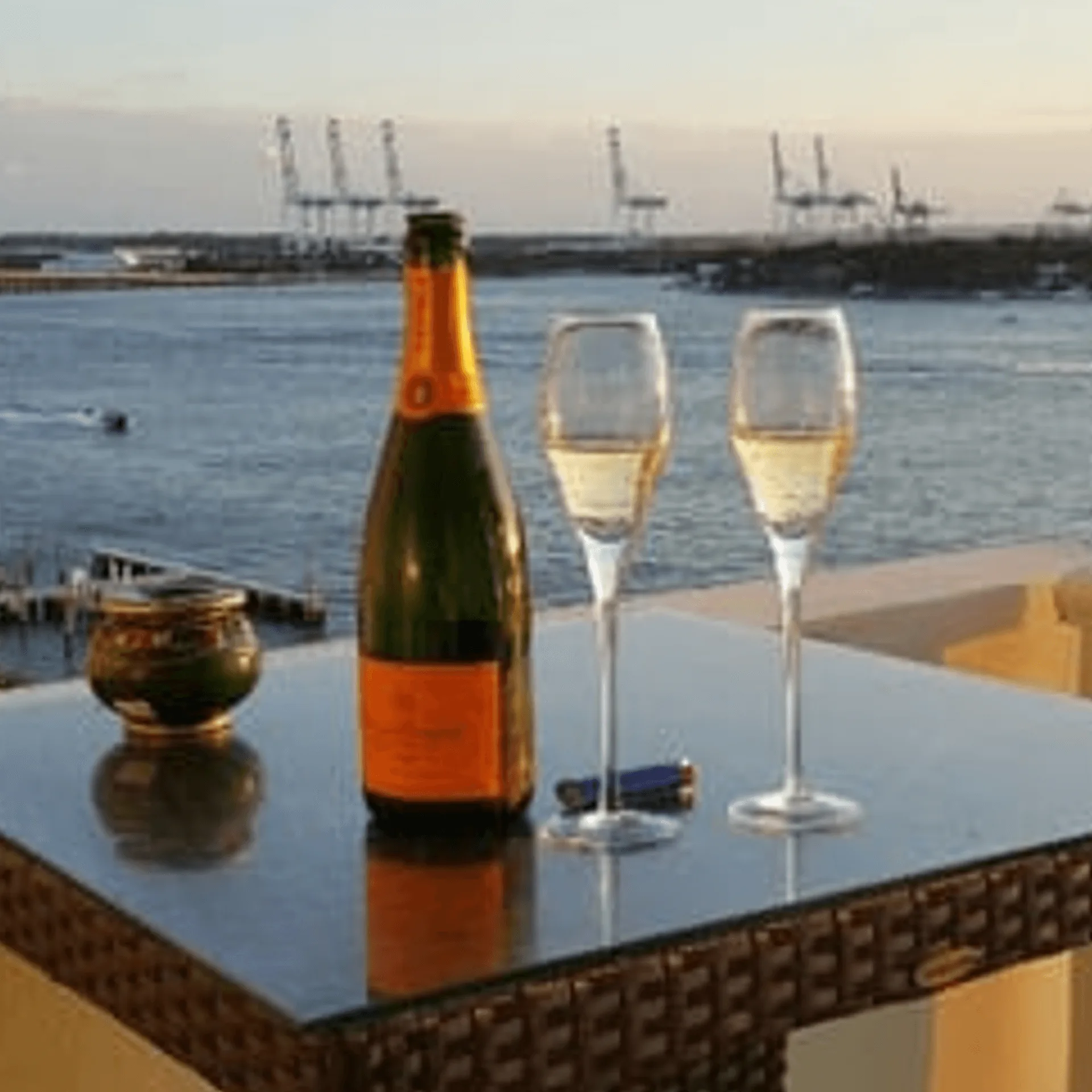 Champagne bottle and two flutes on a table overlooking a harbor with cranes at sunset.