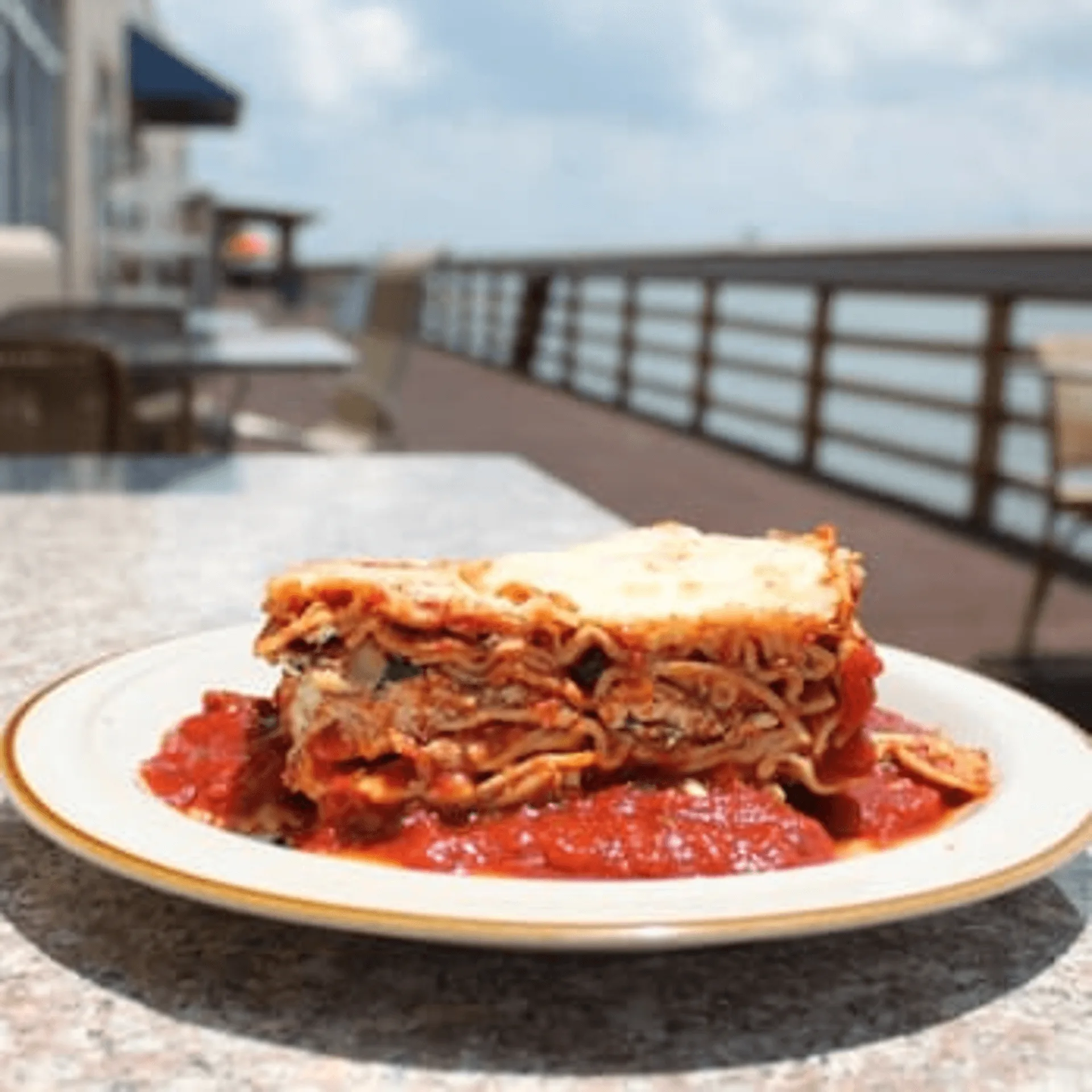 A slice of lasagna with red sauce on a white plate, on an outdoor patio overlooking a pier.