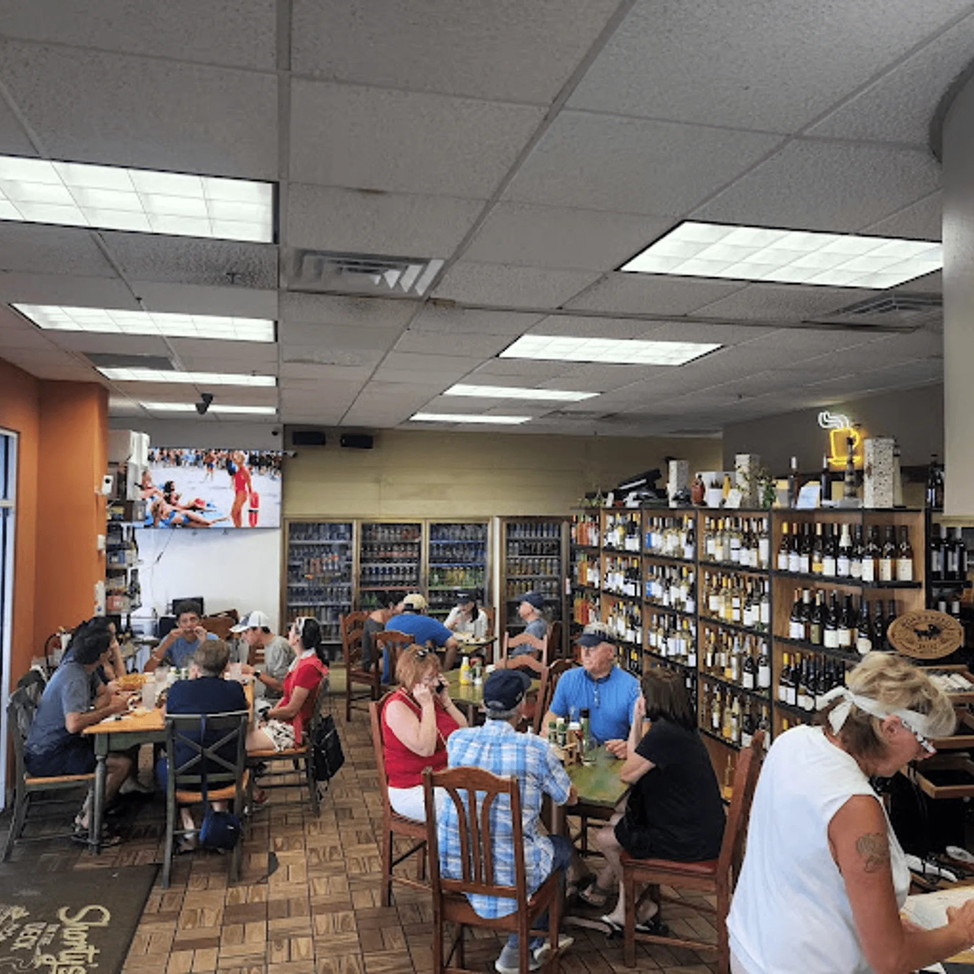Diners seated at tables in a casual restaurant with shelves of wine and beer in the background.