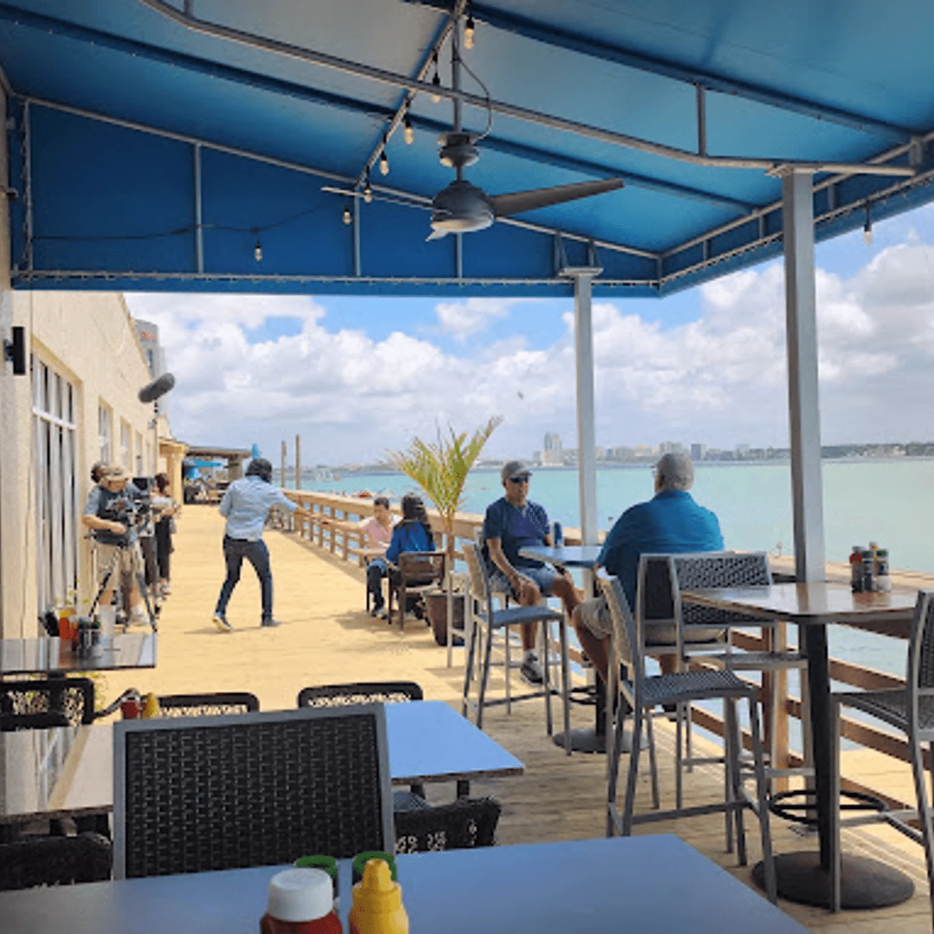 ShortyonDeck's outdoor dining area with people, blue canopy, and water views under a cloudy sky.
