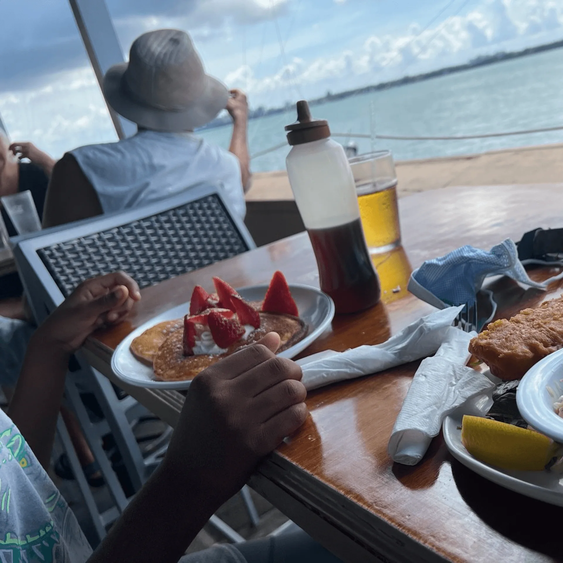 Pancakes with strawberries and whipped cream, beer, and fried fish on a table overlooking the ocean.