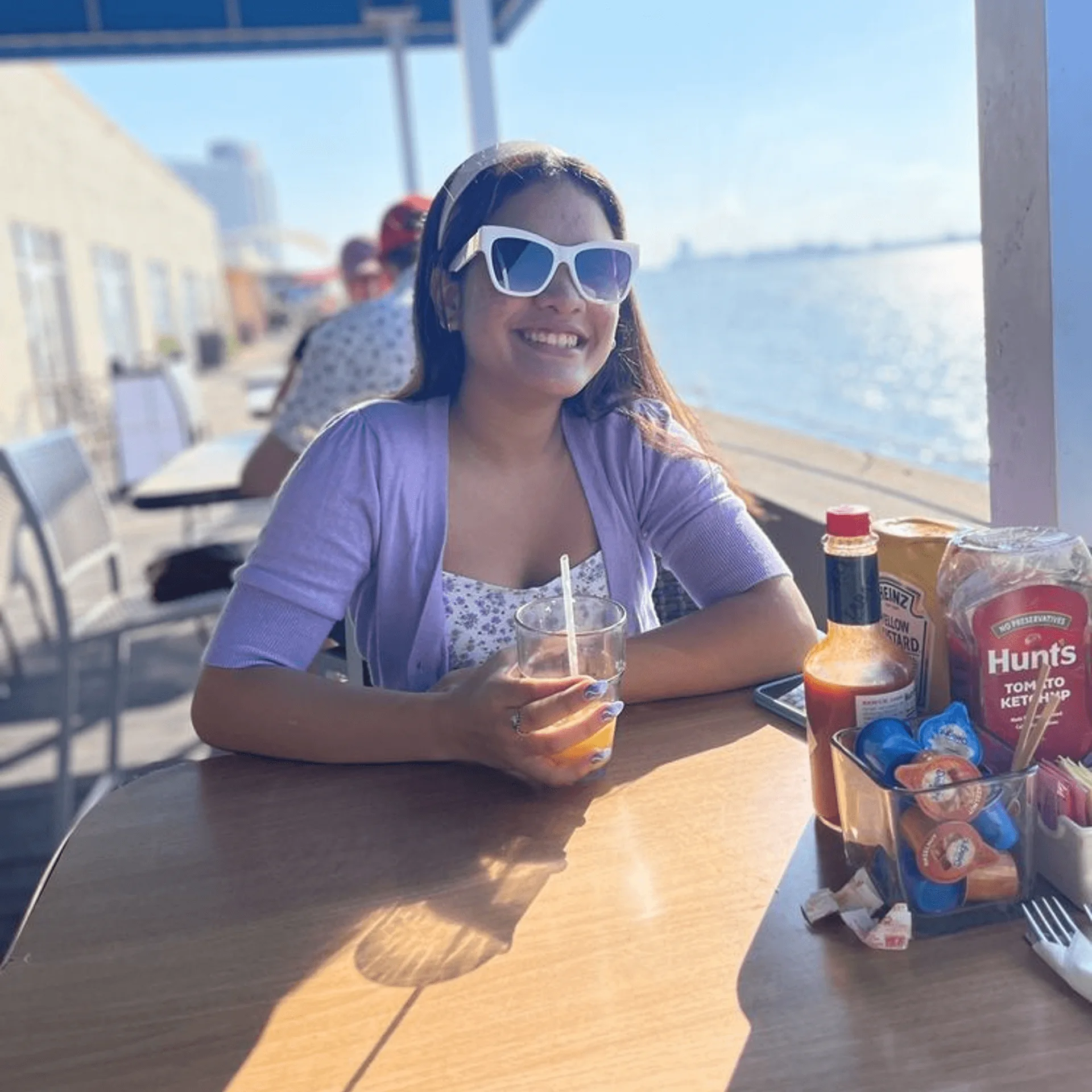 A smiling woman in sunglasses enjoys a drink at an outdoor ShortyonDeck table overlooking the water.